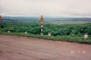 Bức ảnh chụp cảnh quan ven một con đường đất đỏ ở vùng cao nguyên, có thể là một đoạn đường đèo. Dưới góc phải ảnh có dấu ngày tháng "97 7 18".。Ven đường có các cọc tiêu sơn sọc trắng đỏ và hai biển báo giao thông. Biển báo ở giữa là biển cảnh báo nguy hiểm hình tam giác, nền vàng, viền đỏ, với biểu tượng cho thấy một khúc cua gấp sang phải. Biển báo còn lại ở phía xa bên trái cũng có hình dạng tương tự nhưng không rõ biểu tượng.。Con đường không được trải nhựa, có màu đất đỏ đặc trưng. Bên lề đường là các bụi cây xanh rậm rạp. Nhìn ra xa là một khung cảnh thiên nhiên rộng lớn với những ngọn đồi, rừng cây và thung lũng nối tiếp nhau. Bầu trời u ám và đầy mây. Không có người hay hoạt động cụ thể nào được ghi lại, chỉ có phong cảnh thiên nhiên và hạ tầng giao thông đơn sơ của thời kỳ đó.。（日本語訳）。高原地帯にある赤土の道の脇の風景を写した写真で、峠道の一区間かもしれない。写真の右下には「97 7 18」という日付の印がある。道端には赤と白の縞模様に塗られたデリニエーター（視線誘導標）と2つの交通標識がある。中央の標識は、黄色い背景に赤い縁取りの三角形の危険警告標識で、右への急カーブを示すシンボルが付いている。左奥にあるもう一方の標識も同様の形状をしているが、シンボルははっきりしない。道は舗装されておらず、特徴的な赤土の色をしている。道端には緑の茂みが密集している。遠くを見渡すと、丘、森林、谷が連なる広大な自然の風景が広がっている。空はどんよりとしており、雲に覆われている。人や特定の活動は記録されておらず、ただ当時の自然風景と簡素な交通インフラがあるだけである。