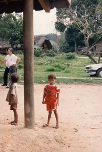 Bối cảnh là một ngôi làng ở vùng nông thôn, có thể là một buôn làng của người dân tộc thiểu số ở khu vực Tây Nguyên. Nền đất là đất đỏ. Ở phía trước, hai bé trai đang đứng chơi. Một bé trai mặc bộ đồ thể thao màu đỏ có chữ 'SPORT' đang nhìn thẳng vào máy ảnh. Bé trai còn lại mặc áo phông và quần đùi, đứng cạnh một cột nhà bằng gỗ lớn. Ở phía sau, có hai người lớn đang đứng, một người đàn ông mặc áo phông trắng và quần jean. Xa hơn là những ngôi nhà sàn truyền thống lợp mái tranh, cây cối xanh tươi và một con lợn đang đi lại trên bãi cỏ. Một phần của một chiếc ô tô màu trắng đậu ở bên phải. Dưới góc trái bức ảnh có dấu ngày '97 7 18'.。（日本語訳）。背景は田舎の村で、おそらくは西部高原地域の少数民族の集落でしょう。地面は赤土です。手前には、二人の男の子が立って遊んでいます。「SPORT」という文字が入った赤いスポーツウェアを着た男の子が、カメラをまっすぐ見ています。もう一人の男の子はTシャツと半ズボンを着て、大きな木製の家の柱の隣に立っています。後ろには二人の大人が立っており、一人の男性は白いTシャツとジーンズを着ています。さらに奥には、茅葺き屋根の伝統的な高床式の家々、青々とした木々があり、一匹の豚が草の上を歩き回っています。右側には白い自動車の一部が停まっています。写真の左下隅には「97 7 18」という日付の印があります。