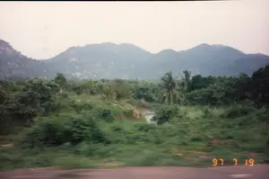 Đây là một bức ảnh phong cảnh được chụp từ một phương tiện đang di chuyển, có thể là ô tô hoặc xe buýt, thể hiện qua phần tiền cảnh bị mờ do chuyển động. Ở góc dưới cùng bên phải của bức ảnh có một dấu ngày tháng kỹ thuật số màu cam ghi '97 7 19'. Khung cảnh là một vùng nông thôn nhiệt đới. Ở phía xa là một dãy núi trập trùng. Gần hơn là một vùng đất với cây cối xanh tươi rậm rạp, bao gồm cả những cây dừa. Một con sông hoặc lạch nhỏ chảy qua khu vực cây cối này. Bức ảnh ghi lại một khoảnh khắc trên một con đường ở vùng quê Việt Nam.。（日本語訳）。これは、動いている乗り物、おそらく自動車かバスから撮影された風景写真で、前景が動きによってぼやけていることからそれがわかります。写真の右下隅には、「'97 7 19」と記されたオレンジ色のデジタル日付スタンプがあります。景色は熱帯の田園地帯です。遠くには、起伏のある山脈があります。もっと手前には、ヤシの木を含む、青々と生い茂る木々のある土地が広がっています。この木々が生い茂る地域を川か小川が流れています。この写真は、ベトナムの田舎道での一瞬を捉えたものです。