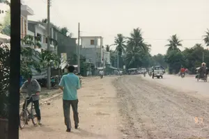 Bức ảnh chụp một khung cảnh nông thôn điển hình của Việt Nam, có lẽ được chụp từ một phương tiện đang di chuyển nên hình ảnh hơi mờ. Ở trung tâm, trên một nền đất cao, có một ngôi nhà tạm nhỏ, đơn sơ. Ngôi nhà có tường vách và mái lợp có thể bằng tôn hoặc lá. Trên nóc nhà có cắm một lá cờ đỏ. Xung quanh ngôi nhà là một khu vườn hoặc cánh đồng với nhiều cây cối xanh tươi, bao gồm cả những cây trông giống cây chuối. Phía sau là quang cảnh rộng lớn của đồng ruộng và cây xanh, xa xa là dãy núi mờ ảo dưới bầu trời u ám. Không có hoạt động nào của con người được ghi lại rõ ràng, chỉ có cảnh vật tĩnh lặng của vùng quê.。（日本語訳）。写真はベトナムの典型的な田舎の風景を写しており、おそらく移動中の乗り物から撮影されたため、画像は少しぼやけています。中央の、少し高くなった土地の上に、小さく簡素な仮設の家があります。家は壁があり、屋根はトタンか葉で葺かれているようです。屋根の上には赤い旗が一本立てられています。後ろには田畑と緑の広大な景色が広がり、遠くには曇り空の下にぼんやりとした山脈が見えます。人間の活動ははっきりと記録されておらず、田舎の静かな風景だけがあります。