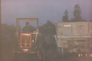 この写真は、夕暮れ時または夜明けに、農村と思われる地域で撮影されたものです。景色はかなり暗く、写真の品質は低いです。左側には赤いトラクターがあり、密閉されたキャビンがないタイプで、運転席に一人が座っています。右側には、トラックまたはトレーラーの後部があります。この二つの車両の間には、集まって立っている人々のグループがおり、おそらく話をしているか、作業の準備をしているようです。光が弱いため、彼らの具体的な活動をはっきりと見ることはできません。後方には、暗い空の下に木々の影があります。背景は、農村地域での農業活動または物資の輸送を示唆しています。写真には文字情報があります。右下隅にはオレンジ色の「\'97 7 19」という日付スタンプがあります。トラックの泥除けには「1M」という文字とその下に数字の列がありますが、はっきりと読み取れません。トラックの側面にも文字が書かれていますが、薄れていて読むことはできません