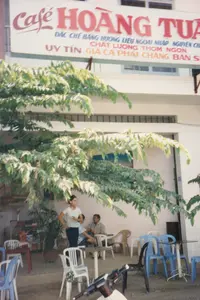 Bức ảnh chụp một quán cà phê ngoài trời ở Việt Nam vào khoảng năm 1997. Phía trên là một tấm biển hiệu lớn của quán có ghi:。"Café HOÀNG TUẤN"。"ĐẶC CHẾ BẰNG HƯƠNG LIỆU NGOẠI NHẬP NGUYÊN CHẤT"。"UY TÍN CHẤT LƯỢNG THƠM NGON"。"GIÁ CẢ PHẢI CHĂNG BÁN SỈ"。Đây là một quán cà phê có không gian mở, có thể là ở vỉa hè hoặc sân trước một tòa nhà. Dưới bóng mát của một cây lớn, có một nhóm khách đang ngồi thư giãn. Một người phụ nữ mặc áo thun trắng đang đứng, và hai người đàn ông đang ngồi tại bàn. Các vật dụng trong ảnh bao gồm bàn và ghế nhựa nhiều màu (trắng, xanh, đen), là loại phổ biến ở các quán nước bình dân thời bấy giờ. Ở phía trước có một phần của một chiếc xe máy. Phía sau là một tòa nhà nhiều tầng.。（日本語訳）。1997年頃のベトナムの屋外カフェを写した写真です。上部には、店の大きな看板があり、次のように書かれています：。「カフェ・ホアン・トゥアン」。「純粋な輸入香料で特別に製造」。「信頼・品質・美味」。「手頃な価格・卸売」。これはオープンスペースのカフェで、歩道か建物の前庭にあるのかもしれません。大きな木の木陰の下で、数人のお客さんが座ってくつろいでいます。白いTシャツを着た女性が一人立っており、二人の男性がテーブルに座っています。写真の中の物には、様々な色（白、青、黒）のプラスチック製のテーブルと椅子が含まれており、これらは当時の庶民的な飲み物屋で一般的なものでした。手前にはバイクの一部が見えます。後ろには多層階の建物があります。