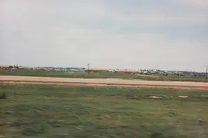 Bức ảnh chụp một khung cảnh vùng nông thôn hoặc ngoại ô, có thể được chụp từ một phương tiện đang di chuyển, thể hiện qua sự mờ nhòe ở tiền cảnh. Tiền cảnh là một bãi cỏ xanh. Ở giữa là một con đường đất màu nâu nhạt chạy ngang qua khung hình. Phía sau con đường là một cánh đồng rộng lớn. Ở phía xa, dọc theo đường chân trời, là một dãy các tòa nhà thấp tầng, có mái màu nâu đỏ, trông giống như nhà ở, nhà kho hoặc một khu nhà tập thể. Có một vài cột điện rải rác trong khung cảnh. Bầu trời có mây và màu sáng. Không có người hay hoạt động cụ thể nào được nhìn thấy. Không có văn bản hay biển báo nào có thể đọc được trong ảnh.。（日本語訳）。写真は田舎または郊外の風景を写したもので、前景のブレからわかるように、移動中の乗り物から撮影された可能性があります。前景は緑の草地です。中央には、フレームを横切る薄茶色の未舗装の道があります。道の後ろには広大な畑が広がっています。遠くの地平線に沿って、赤茶色の屋根を持つ低層の建物が並んでおり、住宅、倉庫、または集合住宅のように見えます。風景の中には電柱が数本点在しています。空は曇っており、明るい色をしています。人や特定の活動は見られません。写真の中には読み取れる文字や標識はありません。