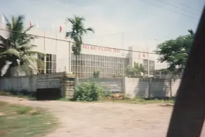 Trên tường của một tòa nhà lớn màu trắng có dòng chữ màu đỏ "NHÀ MÁY BIA RỒNG VÀNG". Đây là một nhà máy bia. Tòa nhà được bao quanh bởi một hàng rào bê tông. Xung quanh có cây cối, bao gồm cả cây dừa và các loại cây xanh khác. Trên nóc nhà có cắm một số lá cờ màu đỏ và trắng. Phía trước tòa nhà là một con đường đất. Bức ảnh dường như được chụp từ một phương tiện đang di chuyển, nhìn ra bên ngoài.。（日本語訳）。大きな白い建物の壁には、「NHÀ MÁY BIA RỒNG VÀNG」という赤い文字があります。これはビール工場です。建物はコンクリートの塀で囲まれています。周りにはヤシの木やその他の緑樹など、木々があります。屋根の上には赤と白の旗が数本立てられています。建物の前は未舗装の道です。写真は移動中の乗り物から外を眺めて撮影されたようです。