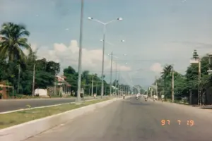 Chữ viết trong ảnh: Dấu ngày tháng màu cam ở góc dưới bên phải ảnh ghi "97 7 19". Không có biển báo hay chữ viết nào khác có thể đọc được.。Mô tả cảnh vật và hoạt động: Bức ảnh chụp một con đường quốc lộ hoặc đại lộ rộng rãi, được trải nhựa ở Việt Nam vào ngày 19 tháng 7 năm 1997. Con đường có dải phân cách cứng ở giữa được trồng cỏ. Dọc theo con đường là một hàng dài các cột đèn đường cao áp hai nhánh. Hai bên đường rợp bóng cây xanh, bao gồm cả những cây dừa, gợi ý về một khung cảnh ở miền Nam hoặc ven biển. Một vài ngôi nhà, trong đó có nhà mái ngói, nằm ven đường. Trên đường, có một số người đang di chuyển bằng xe máy và xe đạp, cho thấy hoạt động giao thông thường ngày nhưng khá thưa thớt. Bầu trời trong xanh, có vài gợn mây trắng.。（日本語訳）。写真の中の文字：写真の右下隅にあるオレンジ色のタイムスタンプには「97 7 19」と記されています。他に読み取れる看板や文字はありません。風景と活動の説明：この写真は、1997年7月19日にベトナムで撮影された、舗装された広々とした国道または大通りを写しています。道路の中央には草が植えられた固定式の中央分離帯があります。道路に沿って、二股の高圧街路灯の柱が長く続いています。道路の両側は緑の木々で覆われており、その中にはココナッツの木も含まれ、南部または沿岸地域の風景であることを示唆しています。瓦屋根の家を含むいくつかの家が道端にあります。道路上では、数人がバイクや自転車で移動しており、日常的でありながらもかなりまばらな交通状況を示しています。空は青く澄んでおり、白い雲がいくつか浮かんでいます。