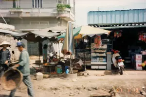 Bức ảnh ghi lại một cảnh sinh hoạt đời thường tại một khu chợ nhỏ ven đường ở Việt Nam, chụp vào ngày 19 tháng 7 năm 1997. Nền đất là đường đất chưa được trải nhựa.。Hoạt động chính là buôn bán tại một khu chợ tự phát. Ở phía trước, một người đàn ông đội mũ lưỡi trai đang đi và mang một chiếc mẹt tre lớn. Bên cạnh ông là một người phụ nữ đội nón lá. Ở trung tâm, nhiều người đang tụ tập quanh các quầy hàng rong được che bằng những chiếc ô lớn và tấm bạt. Một quầy hàng có bày bán các loại hàng hóa trông giống như trái cây.。Bên phải là một cửa hàng tạp hóa có mái tôn, bên trong bày bán nhiều loại hàng hóa đóng gói trên kệ và trong tủ kính. Một chiếc xe máy màu đỏ và trắng được dựng ở phía trước cửa hàng. Có thể thấy một vài thùng carton bên trong, nhưng chữ viết trên đó không rõ ràng, chỉ có thể đoán được chữ "Pro" trên một thùng. Phía sau khu chợ là một tòa nhà bê tông hai tầng.。Dấu ngày tháng "97 7 19" được in ở góc dưới bên phải của bức ảnh.。（日本語訳）。この写真は、1997年7月19日に撮影された、ベトナムの道端にある小さな市場での日常の光景を記録したものです。地面は未舗装の土の道です。主な活動は、自然発生的にできた市場での商売です。手前では、野球帽をかぶった男性が歩いており、大きな竹製の平かごを持っています。彼の隣には、ノンラー（菅笠）をかぶった女性がいます。中央では、多くの人々が大きな傘やシートで覆われた露店の周りに集まっています。右側にはトタン屋根の雑貨店があり、店内では棚やガラスケースに多くの種類の包装された商品が陳列されています。店の前には赤と白のバイクが一台停められています。中にはいくつかの段ボール箱が見えますが、そこに書かれた文字は不鮮明で、一つの箱に「Pro」という文字が推測できるだけです。市場の後ろには、2階建てのコンクリートの建物があります。写真の右下隅には「97 7 19」という日付が印字されています。