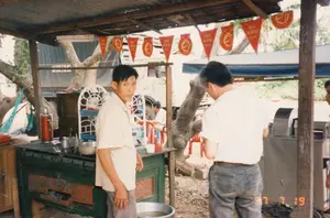 Bối cảnh là một quán nước giải khát ven đường với mái che lợp tôn đơn sơ và nền đất. Có các lá cờ trang trí màu đỏ treo trên mái hiên với dòng chữ 'RED HORSE BEER'. Một người đàn ông, có thể là chủ quán, đang đứng sau quầy hàng bằng gỗ màu xanh. Một khách hàng mặc áo phông trắng đang đứng trước quầy. Ở phía sau, có những chiếc bàn ghế nhựa màu đỏ dành cho khách và một người đàn ông khác đang ngồi. Bên trái có một người đang nằm nghỉ trên võng. Trên quầy có các vật dụng như phích nước màu đỏ và bát kim loại. Nổi bật ở bên phải là một chiếc máy ép nước mía, cho thấy đây là nơi bán nước mía. Phía sau quán có một chiếc xe tải đang đỗ. Khung cảnh này mô tả một điểm dừng chân nghỉ ngơi phổ biến dọc các tuyến đường ở Việt Nam vào thời điểm đó.。（日本語訳）。背景は、簡素なトタン葺きの屋根と土の地面を持つ、道端の飲料店です。軒先には「RED HORSE BEER」という文字が入った赤い装飾用の旗が吊るされています。店主と思われる男性が、青い木製の屋台カウンターの後ろに立っています。白いTシャツを着た客が一人、カウンターの前に立っています。後方には、客用の赤いプラスチック製のテーブルと椅子があり、別の男性が一人座っています。左側には、ハンモックで横になって休んでいる人が一人います。カウンターの上には、赤い魔法瓶や金属製のボウルなどの物があります。右側で目立っているのはサトウキビ搾り機で、ここがサトウキビジュースを売る場所であることを示しています。店の後ろにはトラックが一台駐車しています。この光景は、当時のベトナムの道路沿いによく見られた休憩所を描写しています。