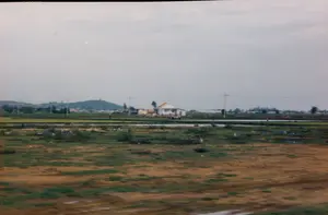 Bức ảnh chụp một khung cảnh nông thôn ở Việt Nam, có thể là trên tuyến đường giữa Nha Trang và Sài Gòn. Cảnh vật chủ yếu là những cánh đồng lúa rộng lớn. Ở giữa bức ảnh, có một ngôi nhà một tầng, tường trắng và mái ngói màu cam đỏ. Xung quanh ngôi nhà là những thửa ruộng lúa, một số đã được cấy lúa xanh, một số khác vẫn còn ngập nước. Có thể thấy một vài người nông dân đang làm việc trên đồng, có lẽ họ đang cấy lúa hoặc chăm sóc cây trồng. Phía sau, xa xa là những ngọn đồi và đường dây điện chạy ngang qua khung cảnh. Bầu trời có nhiều mây. Phần tiền cảnh của bức ảnh bị mờ, cho thấy nó có thể được chụp từ một phương tiện đang di chuyển. Không có văn bản hay chữ viết nào có thể đọc được trong ảnh.。（日本語訳）。写真はベトナムの田園風景を写したもので、おそらくニャチャンとサイゴンの間の路上で撮影されたものです。風景は主に広大な水田です。写真の中央には、白い壁と赤みがかったオレンジ色の瓦屋根を持つ一階建ての家があります。家の周りには水田が広がっており、一部には青い稲が植えられ、他の部分はまだ水に浸かっています。数人の農民が畑で作業しているのが見えます。おそらく田植えや作物の手入れをしているのでしょう。後方、遠くには丘があり、電線が風景を横切っています。空は雲が多いです。写真の前景はぼやけており、移動中の乗り物から撮影された可能性を示しています。写真には読み取れるテキストや文字はありません。