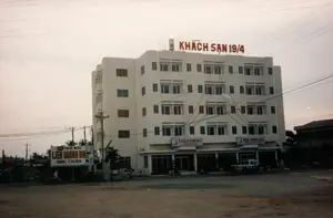 Bản ghi chép các văn bản trong hình ảnh:。Trên nóc tòa nhà: "KHÁCH SẠN 19/4".。Trên các biển hiệu ở tầng trệt của tòa nhà: "KHÁCH SẠN 19/4 Hotel" và "NHÀ HÀNG 19/4 Restaurant".。Trên tấm biển quảng cáo lớn bên trái: "BGI NHA MAY LIÊN DOANH BIA BÌNH THUẬN".。Mô tả cảnh quan và hoạt động:。Bức ảnh chụp một tòa nhà lớn, nhiều tầng màu trắng, là "Khách sạn 19/4". Tòa nhà có kiến trúc khá hiện đại so với thời điểm năm 1997. Tầng trệt của tòa nhà là khu vực nhà hàng và sảnh khách sạn. Phía trước khách sạn là một khoảng đất rộng, không được trải nhựa, có thể là bãi đậu xe hoặc một khu đất trống. Một chiếc xe ô tô địa hình màu trắng đang đỗ ở phía trước nhà hàng.。Bên trái của tòa nhà có một tấm biển quảng cáo lớn cho "Nhà máy Liên doanh Bia Bình Thuận". Gần tấm biển này có một vài chiếc xe máy đang đỗ và một người đang ngồi. Bầu trời u ám, và có rất nhiều dây điện, dây cáp chằng chịt phía trên tòa nhà và đường phố, một hình ảnh quen thuộc của các đô thị Việt Nam thời kỳ này. Cảnh vật xung quanh cho thấy đây là một khu vực đang trong quá trình phát triển.。（日本語訳）。画像内のテキストの記録：。建物の屋上：「KHÁCH SẠN 19/4」。建物の1階にある看板：「KHÁCH SẠN 19/4 Hotel」と「NHÀ HÀNG 19/4 Restaurant」。左側の大きな広告看板：「BGI NHA MAY LIÊN DOANH BIA BÌNH THUẬN」。風景と活動の説明：。写真は、「4月19日ホテル」である、白くて大きな多層階の建物を写しています。建物は1997年当時としてはかなり近代的な建築様式です。建物の1階はレストランとホテルのロビーエリアです。ホテルの前には舗装されていない広い土地があり、駐車場か空き地である可能性があります。レストランの前に白いオフロード車が1台駐車しています。建物の左側には、「ビン・トゥアンビール合弁工場」の大きな広告看板があります。この看板の近くには数台のバイクが駐車しており、一人の人が座っています。空は曇っており、建物や通りの上にはたくさんの電線やケーブルが張り巡らされています。これはこの時代のベトナムの都市部ではおなじみの光景です。周囲の風景は、ここが開発途上にある地域であることを示しています。