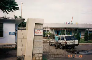Bức ảnh chụp lối vào của một cơ quan nhà nước vào ngày 19 tháng 7 năm 1997.。Chữ viết trên các bảng hiệu:。1. Bảng hiệu chính gắn trên cột cổng lớn bằng đá ghi: "SỞ GIAO THÔNG VẬN TẢI BÌNH THUẬN".。2. Một bảng thông báo nhỏ hơn màu xanh lam gắn trên tường của tòa nhà bên trái có dòng chữ "YÊU CẦU" ở trên cùng, các dòng chữ còn lại ở dưới bị mờ, không thể đọc rõ toàn bộ nội dung, dường như là quy định cho khách đến làm việc.。Mô tả cảnh vật và hoạt động:。Đây là khuôn viên của một văn phòng chính phủ, cụ thể là Sở Giao thông Vận tải tỉnh Bình Thuận. Một tòa nhà văn phòng một tầng nằm ở phía sau, trên nóc nhà có treo một lá cờ đỏ và một dãy cờ đuôi nheo nhiều màu sắc.。Bên trong cổng sắt, một chiếc ô tô kiểu station wagon màu trắng đang đỗ. Một người đàn ông mặc áo sơ mi màu xanh nhạt đang đứng cạnh chiếc xe, hai tay đưa lên đầu. Một người đàn ông khác đang đi bộ trong sân ở phía sau. Cổng vào được làm bằng sắt và có một cột trụ lớn bằng đá. Nền đất có vài chỗ còn đọng nước, cho thấy trời có thể vừa mưa.。（日本語訳）。1997年7月19日に撮影された、ある政府機関の入り口の写真。看板の文字：。1. 大きな石造りの門柱に取り付けられたメインの看板には、「SỞ GIAO THÔNG VẬN TẢI BÌNH THUẬN」（ビントゥアン省交通運輸局）と書かれている。2. 左側の建物の壁に取り付けられた青色の小さな案内板には、上部に「YÊU CẦU」（要求）という文字があり、その下の残りの文字はぼやけていて、内容全体をはっきりと読み取ることはできないが、来訪者向けの規定のようである。風景と活動の説明：。これは政府機関、具体的にはビントゥアン省交通運輸局の敷地である。鉄製の門の内側には、白いステーションワゴン型の自動車が1台駐車している。水色のシャツを着た男性が車の横に立っており、両手を頭の上に上げている。別の男性が後方の中庭を歩いている。入り口の門は鉄製で、大きな石の柱がある。地面には水たまりが数か所残っており、雨が降ったばかりかもしれないことを示している。