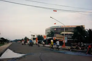 Khung cảnh một con đường lớn ở một đô thị Việt Nam vào khoảng năm 1997. Trên một tòa nhà lớn, hiện đại có dòng chữ 'NGÂN HÀNG CÔNG THƯƠNG VIỆT NAM - CHI NHÁNH...' và 'INDUSTRIAL AND COMMERCIAL BANK OF VIET NAM - ... BRANCH'. Phần tên chi nhánh bị cây che khuất. Trên nóc tòa nhà có cờ Việt Nam. Treo vắt ngang đường phía trên là một biểu ngữ màu đỏ ghi 'ĐƯỜNG DÂY ĐIỆN THOẠI' và 'ĐỘ CAO 5m'.。Trên đường, người dân đang di chuyển bằng các phương tiện phổ biến thời bấy giờ như xe đạp, xe máy và xích lô. Có một người phụ nữ đội nón lá, mặc bộ đồ màu vàng chấm bi đang đạp xe. Bên cạnh các tòa nhà là những hàng quán nhỏ, lợp mái tôn. Bên lề đường, một nhóm người đang tụ tập buôn bán nhỏ, ngồi trên những chiếc ghế đẩu. Dọc hai bên đường có nhiều cây xanh, cột đèn và treo nhiều cờ, biểu ngữ màu đỏ.。（日本語訳）。1997年頃のベトナムのある都市の大きな通りの光景。大きく近代的な建物には、「NGÂN HÀNG CÔNG THƯƠNG VIỆT NAM - CHI NHÁNH...」と「INDUSTRIAL AND COMMERCIAL BANK OF VIET NAM - ... BRANCH」の文字がある。支店名の部分は木で隠れている。建物の屋上にはベトナム国旗がある。路上では、人々が自転車、バイク、シクロといった当時の一般的な乗り物で移動している。ノンラーをかぶり、水玉模様の黄色い服を着た女性が自転車をこいでいる。建物の隣には、トタン屋根の小さな店が並んでいる。道端では、一群の人々が小さなスツールに座り、集まって小商いをしている。