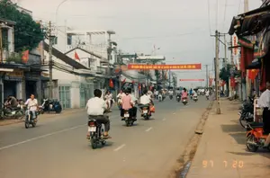 Chữ viết trên các biểu ngữ và biển hiệu:。1. Biểu ngữ lớn màu đỏ giăng ngang đường: "BỎ PHIẾU BẦU CỬ ĐẠI BIỂU QUỐC HỘI LÀ QUYỀN VÀ NGHĨA VỤ CỦA MỖI CÔNG DÂN".。2. Biển hiệu cửa hàng bên trái, chữ màu đỏ trên nền vàng: "HUY HOANG".。3. Biển hiệu dọc bên phải: "HẠNH PHÚC".。4. Dấu ngày tháng ở góc dưới bên phải: "97 7 20".。Mô tả cảnh vật và hoạt động:。Đây là khung cảnh trên một con đường phố ở Việt Nam vào ngày 20 tháng 7 năm 1997. Con đường được trải nhựa với vạch kẻ đường, hai bên là vỉa hè và các ngôi nhà phố san sát nhau. Các ngôi nhà này có kiến trúc dạng ống, cao từ 2 đến 3 tầng, vừa để ở vừa làm mặt bằng kinh doanh.。Hoạt động chính là giao thông tấp nập với rất nhiều người đang điều khiển xe máy (xe gắn máy) trên đường. Xe máy là phương tiện di chuyển chủ yếu, có cả người lái một mình và người chở thêm người ngồi sau. Một số xe máy khác đang đậu trên vỉa hè. Không khí trên đường phố cho thấy một ngày sinh hoạt bình thường, tuy nhiên, việc treo rất nhiều cờ đỏ sao vàng và biểu ngữ tuyên truyền về bầu cử cho thấy đây là một ngày có sự kiện chính trị quan trọng. Dựa vào nội dung biểu ngữ và ngày tháng trên ảnh, đây chính là ngày diễn ra cuộc bầu cử Đại biểu Quốc hội.。Các vật thể nổi bật bao gồm: rất nhiều xe máy các loại (phổ biến là dòng Honda Cub), các cột điện bằng bê tông với hệ thống dây điện và dây cáp chằng chịt, các biển hiệu cửa hàng, cờ tổ quốc và biểu ngữ chính trị.。（日本語訳）。横断幕と看板の文字：。1. 道路を横切る大きな赤い横断幕：「国会議員選挙の投票は、すべての国民の権利であり義務です」。2. 左側の店の看板、黄色地に赤文字：「HUY HOANG」。3. 右側の縦看板：「HẠNH PHÚC」。4. 右下隅の日付スタンプ：「97 7 20」。風景と活動の描写：。これは1997年7月20日のベトナムのある街頭の光景です。道路は車線が引かれたアスファルト舗装で、両側には歩道と商店が密集しています。これらの家は筒状の建築様式で、2階から3階建てで、住居と商業スペースを兼ねています。主な活動は、多くの人々が道路でバイク（原動機付自転車）を運転している、にぎやかな交通です。バイクが主要な移動手段であり、一人で運転する人もいれば、後ろに人を乗せている人もいます。他のいくつかのバイクは歩道に駐車されています。横断幕の内容と写真の日付に基づくと、これはまさに国会議員選挙が行われる日です。目立つ物体には、多くの種類のバイク（ホンダカブシリーズが一般的）、複雑に絡み合った電線やケーブルがあるコンクリート製の電柱、店の看板、国旗、そして政治的な横断幕が含まれます。