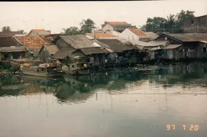 Bức ảnh ghi lại khung cảnh một khu dân cư ven sông hoặc kênh rạch vào ngày 20 tháng 7 năm 1997. Dọc theo bờ là một dãy nhà san sát nhau, phần lớn là các ngôi nhà sàn được dựng trên mặt nước bằng các vật liệu đơn sơ như gỗ, với mái lợp tôn, mái lá, xen kẽ với một vài ngôi nhà xây kiên cố hơn lợp ngói. Trên các mái nhà có rất nhiều ăng-ten TV. Dưới mặt nước có hai chiếc ghe đang neo đậu. Một chiếc ghe lớn hơn có người đang ngồi trên đó, dường như đang sinh hoạt hoặc nghỉ ngơi, trên ghe có chở các vật dụng như những chiếc lu sành lớn. Chiếc ghe còn lại nhỏ hơn. Mặt nước kênh có màu đục, phản chiếu bóng của những ngôi nhà và cây cối. Trên một tòa nhà ở phía xa có một bảng hiệu với chữ 'TUẤN'. Góc dưới cùng bên phải của ảnh có dấu ngày kỹ thuật số '97 7 20'.。（日本語訳）。写真は1997年7月20日の、川または運河沿いのある住宅地の風景を記録したものです。岸に沿って家々が密集して並んでおり、その大部分は木材のような簡素な材料で水上に建てられた高床式の家で、トタン屋根、葉葺き屋根であり、その間に瓦葺きのより頑丈な家がいくつか混ざっています。屋根の上にはたくさんのテレビアンテナがあります。水面には2艘の小舟が停泊しています。大きい方の小舟には人が乗っており、生活しているか、あるいは休憩しているようで、舟には大きな陶器の壺のような物が積まれています。残りの小舟はより小さいです。運河の水面は濁っており、家々や木々の影を映しています。遠くの建物には「TUẤN」という文字の看板があります。写真の右下隅には「'97 7 20」というデジタルの日付スタンプがあります。