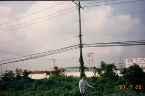 Bản ghi văn bản từ hình ảnh:。Trên tấm biển quảng cáo lớn màu trắng có ghi:。- KHU CHẾ XUẤT LINH TRUNG。- LINH TRUNG EXPORT PROCESSING ZONE。- 領中加工出口區 (chữ Hán)。Phía dưới có các dòng chữ nhỏ hơn nhưng không thể đọc rõ, có thể là thông tin liên lạc.。Trên ảnh có dấu ngày tháng màu vàng: '97 7 20'.。Mô tả bối cảnh và hoạt động:。Bức ảnh được chụp vào ngày 20 tháng 7 năm 1997, tại một khu vực ngoại ô, có thể là gần Sài Gòn.。- **Loại địa điểm:** Đây là một khu đất trống, hoang sơ với cây cỏ mọc um tùm, nằm ngay cạnh hàng rào của một khu công nghiệp hoặc khu chế xuất. Tấm biển quảng cáo xác nhận đây là khu vực của "Khu Chế Xuất Linh Trung".。- **Hoạt động:** Một thiếu niên mặc áo sơ mi dài tay sáng màu và quần tối màu đang đứng giữa đám cây cối. Không có hoạt động cụ thể nào đang diễn ra, dường như đây là một khoảnh khắc được chụp lại để ghi lại cảnh quan.。- **Các vật thể trong ảnh:**。- Một thiếu niên.。- Một tấm biển quảng cáo lớn giới thiệu về Khu Chế Xuất Linh Trung.。- Nhiều cột điện bê tông với hệ thống dây điện chằng chịt bắc ngang qua bầu trời, một đặc điểm phổ biến của cảnh quan đô thị và ven đô Việt Nam thời kỳ này. Một trong những cột điện ở gần nhất có dây leo quấn quanh thân.。- Ở phía sau là các tòa nhà nhà xưởng và tường rào của khu công nghiệp.。- Bầu trời có mây.。（日本語訳）。画像からの文字起こし：。大きな白い看板には次のように書かれています：。- KHU CHẾ XUẤT LINH TRUNG。- LINH TRUNG EXPORT PROCESSING ZONE。- 領中加工出口區 (漢字)。下部にはより小さな文字の行がありますが、はっきりと読み取ることはできず、連絡先情報である可能性があります。写真には黄色い日付スタンプ「'97 7 20」があります。背景と活動の説明：。写真は1997年7月20日に、サイゴン近郊と思われる郊外の地域で撮影されました。- **場所の種類：** これは、草木が生い茂る未開拓の空き地で、工業団地または輸出加工区のフェンスのすぐ隣に位置しています。看板は、ここが「リンチュン輸出加工区」の区域であることを示しています。- **活動：** 明るい色の長袖シャツと暗い色のズボンを着用した一人の少年が木々の中に立っています。特定の活動は行われておらず、風景を記録するために撮影された一瞬のようです。- **写真の中の物体：**。- 一人の少年。- リンチュン輸出加工区を紹介する大きな看板。- 多数のコンクリート製電柱と、空を横切る複雑に絡み合った電線システム。これはこの時期のベトナムの都市部および郊外の景観に共通する特徴です。最も近くにある電柱の一つには、蔓が巻き付いています。- 後方には、工業団地の工場建物と塀があります。- 空には雲があります。