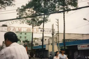 Bức ảnh ghi lại một khung cảnh đường phố ở Sài Gòn vào năm 1997.。**Thông tin chữ viết:**。*   Một cổng chào lớn bằng kim loại bắc ngang qua đường có các dòng chữ: "CÔNG VIÊN VĂN HÓA" ở trên cùng và "ĐẦM SEN" là dòng chữ lớn nhất ở giữa. Bên cạnh chữ "ĐẦM SEN" có các ký tự chữ Hán "文化公園" (Văn Hóa Công Viên) và "蓮潭" (Liên Đàm).。*   Ở phía bên phải, có một biển hiệu của một cửa hàng ghi: "CỬA HÀNG XE GẮN MÁY", và dòng chữ nhỏ hơn bên dưới có thể là "MUA BÁN TRAO ĐỔI KÝ GỞI CÁC LOẠI XE GẮN MÁY".。*   Có những biển hiệu khác trên các tòa nhà nhưng không thể đọc rõ, ví dụ như một biển hiệu có chữ "Quỳnh Hương" bị mờ.。**Mô tả cảnh vật và hoạt động:**。*   **Loại địa điểm:** Đây là một con đường đô thị, cụ thể là lối vào Công viên Văn hóa Đầm Sen. Hai bên đường là các tòa nhà phố nhiều tầng liền kề được sử dụng làm nhà ở và cửa hàng kinh doanh.。*   **Hoạt động:** Cảnh tượng thể hiện hoạt động giao thông thường ngày. Một người đàn ông mặc áo sơ mi sọc ở tiền cảnh dường như đang ngồi trên xe máy. Ở phía sau, nhiều người khác cũng đang di chuyển trên đường bằng xe máy và đi bộ.。*   **Vật thể:** Các vật thể nổi bật bao gồm cổng chào lớn, các tòa nhà, nhiều cây xanh cao lớn, cột điện, hệ thống dây điện và dây cáp chằng chịt, đèn đường và xe gắn máy. Bầu trời có mây, ánh sáng dịu.。（日本語訳）。この写真は1997年のサイゴンのある街の風景を捉えたものです。**書かれている文字の情報：**。*   道路をまたぐ大きな金属製の歓迎ゲートには、一番上に「CÔNG VIÊN VĂN HÓA」（文化公園）、中央に最も大きな文字で「ĐẦM SEN」（ダムセン）と書かれています。「ĐẦM SEN」の文字の隣には、漢字の「文化公園」（Văn Hóa Công Viên）と「蓮潭」（Liên Đàm）があります。*   右側には、ある店の看板に「CỬA HÀNG XE GẮN MÁY」（バイク店）と書かれており、その下の小さな文字は「MUA BÁN TRAO ĐỔI KÝ GỞI CÁC LOẠI XE GẮN MÁY」（各種バイクの売買・交換・委託販売）だと思われます。例えば、「Quỳnh Hương」という文字が書かれたぼやけた看板があります。**風景と活動の描写：**。*   **場所の種類：** これは都市の道路で、具体的にはダムセン文化公園への入り口です。道路の両側には、住居や店舗として利用されている隣接した多層階のタウンハウスが並んでいます。*   **活動：** この光景は日常の交通活動を表しています。前景には、縞模様のシャツを着た男性がバイクに座っているように見えます。後方では、他の多くの人々もバイクや徒歩で道路を移動しています。*   **物体：** 目立つ物体には、大きな歓迎ゲート、建物、多くの背の高い緑の木々、電柱、複雑に絡み合った電線とケーブル、街灯、バイクなどがあります。空は曇っており、光は柔らかいです。
