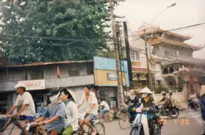 Bức ảnh ghi lại cảnh sinh hoạt trên một con đường ở Việt Nam, vào ngày 20 tháng 7 năm 1997. Con đường khá đông đúc với nhiều người đang di chuyển bằng xe đạp và xe máy.。Về hoạt động và con người, dòng người đang tham gia giao thông. Ở tiền cảnh, có nhiều người đi xe đạp và xe máy. Một người đàn ông đội mũ lưỡi trai trắng đang đạp xe. Một người khác đi xe máy đeo khẩu trang. Một phụ nữ đội nón lá ngồi sau xe. Phía sau họ, nhiều người khác cũng đang đi xe, trong đó có một phụ nữ khác đội nón lá và đeo khẩu trang đang điều khiển một chiếc xe máy màu xanh lam. Việc đội nón lá và đeo khẩu trang cho thấy đây là một ngày nắng và cũng là thói quen để chống bụi khi đi đường.。Về địa điểm và vật thể, hai bên đường là các dãy nhà phố. Có một ngôi nhà cũ với mái tôn, phía trên có treo một lá cờ đỏ. Nổi bật ở phía sau là một công trình kiến trúc lớn, có mái ngói nhiều tầng cong vút theo kiểu truyền thống Á Đông, có khả năng là một ngôi chùa hoặc đình. Hệ thống cột điện và dây điện chằng chịt vắt ngang qua khung hình. Trên một ngôi nhà có một tấm biển hiệu màu trắng, chữ đỏ, có thể đọc được dòng chữ "TƯƠNG NAM", các chữ còn lại trên biển hiệu không rõ. Các phương tiện trong ảnh chủ yếu là xe đạp và các loại xe máy phổ thông thời kỳ đó.。（日本語訳）。この写真は1997年7月20日、ベトナムのある道路での生活風景を記録したものです。道路はかなり混雑しており、多くの人々が自転車やバイクで移動しています。人々の活動については、多くの人が交通に参加しています。前景には、自転車やバイクに乗る多くの人々がいます。白い野球帽をかぶった男性が自転車を漕いでいます。別の人はバイクに乗り、マスクを着用しています。ノンラーをかぶった女性がバイクの後ろに乗っています。彼らの後ろにも、多くの人々が乗り物に乗っており、その中にはノンラーをかぶりマスクを着用して青いバイクを運転している別の女性もいます。ノンラーをかぶりマスクを着用していることは、この日が晴れの日であること、そして道路を移動する際の埃を防ぐための習慣であることを示しています。場所と物については、道路の両側には商店が立ち並んでいます。トタン屋根の古い家があり、その上には赤い旗が掲げられています。後方に目立つのは、東アジアの伝統的な様式で反り返った多層の瓦屋根を持つ大きな建築物で、寺院か村の集会所（ディン）である可能性があります。電柱と電線のシステムが複雑に絡み合い、フレームを横切っています。ある家には白地に赤文字の看板があり、「TƯƠNG NAM」という文字が読み取れますが、看板の残りの文字は不明瞭です。写真の中の乗り物は、主に自転車と当時の一般的な種類のバイクです。