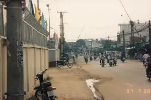 Đây là hình ảnh chụp một con đường ở Việt Nam vào ngày 20 tháng 7 năm 1997. Con đường nhựa có nhiều người đang tham gia giao thông, chủ yếu bằng xe máy, ngoài ra còn có một người đi xe đạp. Phía xa có một chiếc xe tải nhỏ màu trắng. Lề đường bên trái là đường đất, có một vũng nước đọng. Dọc theo lề đường là một bức tường bê tông, phía trên có hàng rào lưới thép. Một dãy cờ nhiều màu sắc (đỏ, vàng, xanh lam, xanh lá) được treo dọc theo hàng rào, có thể là để trang trí cho một ngày lễ hoặc sự kiện. Phía sau là cảnh nhà cửa, cây cối, cùng với hệ thống cột điện và dây điện chằng chịt. Một người đi bộ ở phía xa đang đội nón lá. Trên cột điện ở tiền cảnh có dán giấy quảng cáo đã rách, không đọc được rõ nội dung. Một chiếc xe máy hiệu Honda đang đậu ở lề đường.。（日本語訳）。これは1997年7月20日にベトナムのある道路で撮影された写真です。舗装された道路には多くの人々が交通に参加しており、主にバイクですが、その他に自転車に乗っている人も一人います。遠くには白い小型トラックが一台あります。左側の路肩は未舗装で、水たまりができています。路肩に沿ってコンクリートの壁があり、その上には金網フェンスがあります。フェンスに沿って色とりどり（赤、黄、青、緑）の旗が並んで掲げられており、祝日やイベントの装飾かもしれません。後方には家々や木々、そして複雑に絡み合った電柱と電線のシステムが見えます。遠くで歩いている人が一人、ノンラー（円錐形の葉笠）をかぶっています。前景の電柱には破れた広告紙が貼られていますが、内容ははっきりと読み取れません。ホンダ製のバイクが一台、路肩に停まっています。