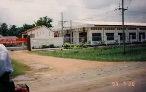 Bức ảnh chụp quang cảnh bên ngoài cổng một xí nghiệp ở Long An vào ngày 20 tháng 7 năm 1997.。**Thông tin chữ viết:**。*   Trên tấm biển hiệu lớn xây bằng gạch ở cổng có ghi: "XÍ NGHIỆP MAY XK LONG AN" (Xí nghiệp May Xuất khẩu Long An). Có một số dòng chữ nhỏ hơn bên dưới nhưng không thể đọc rõ.。*   Một băng rôn lớn màu đỏ được căng ngang cổng vào với nội dung: "NHIỆT LIỆT CHÀO MỪNG ĐẠI BIỂU QUỐC HỘI KHÓA X (20-7-1997)".。**Mô tả cảnh vật và hoạt động:**。*   **Loại địa điểm:** Đây là cổng vào của một nhà máy sản xuất, cụ thể là một xí nghiệp may mặc để xuất khẩu. Tòa nhà chính là một công trình một tầng, sơn trắng, có kiến trúc kiểu nhà xưởng công nghiệp với các ô thông gió phía trên cửa sổ. Toàn bộ khuôn viên được rào lại cẩn thận.。*   **Hoạt động:** Bối cảnh cho thấy xí nghiệp đang chuẩn bị hoặc đang diễn ra một sự kiện quan trọng là đón tiếp các đại biểu Quốc hội. Điều này được thể hiện qua băng rôn chào mừng và các lá cờ phướn nhỏ, nhiều màu sắc được treo dọc theo hàng rào và tòa nhà. Ở phía trước, có một người đang ngồi trên một phương tiện (có thể là xe máy hoặc một loại xe 4 bánh nhỏ) và bức ảnh dường như được chụp từ phương tiện này khi đang di chuyển.。*   **Các vật thể:**。*   Tòa nhà nhà xưởng một tầng.。*   Cổng sắt và hàng rào bao quanh.。*   Biển hiệu xí nghiệp và băng rôn chào mừng.。*   Cột điện, đường dây điện và một trạm biến áp nhỏ.。*   Phía sau là cây cối, bao gồm cả cây dừa, đặc trưng cho vùng nông thôn. Lề đường là đường đất và cỏ mọc.。（日本語訳）。1997年7月20日、ロンアン省にある企業の門の外の光景を撮影した写真。**文字情報：**。*   門にあるレンガ造りの大きな看板には、「XÍ NGHIỆP MAY XK LONG AN」（ロンアン縫製輸出企業）と書かれている。その下にはいくつかの小さな文字の行があるが、はっきりと読むことはできない。*   大きな赤い横断幕が入り口の門に渡って張られており、その内容は「第10期国会議員を熱烈に歓迎します（1997年7月20日）」である。**風景と活動の説明：**。*   **場所の種類：** これは生産工場の入り口であり、具体的には輸出用の縫製企業である。主要な建物は平屋建てで白く塗られ、窓の上部に換気口がある工業用工場の様式の建築である。敷地全体は慎重に柵で囲まれている。*   **活動：** 背景は、企業が国会議員団を迎えるという重要なイベントを準備中または開催中であることを示している。このことは、歓迎の横断幕や、柵と建物に沿って掲げられた小さく色とりどりののぼり旗によって示されている。前方には、一人の人が乗り物（バイクか小型の四輪車の可能性がある）に乗っており、写真はこの乗り物から移動中に撮影されたようである。*   **物体：**。*   平屋建ての工場。*   鉄の門と周囲の柵。*   企業の看板と歓迎の横断幕。*   電柱、電線、そして小さな変電所。*   後方には、田舎の地域に特徴的なヤシの木を含む木々がある。道端は未舗装で草が生えている。