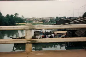 Bức ảnh được chụp từ trên một cây cầu, nhìn xuống một con sông hoặc kênh rạch. Ở phía trước là lan can bê tông của cây cầu. Dưới mặt nước, có một chiếc ghe lớn, dường như là một ngôi nhà nổi, nơi người dân sinh sống. Trên ghe có một dây phơi đầy quần áo nhiều màu sắc đang được phơi khô, thể hiện một cảnh sinh hoạt đời thường. Dọc theo bờ sông là những ngôi nhà, bao gồm cả những ngôi nhà kiên cố ở phía xa và những ngôi nhà lợp mái tôn hoặc mái lá tạm bợ ở gần bờ. Trên các mái nhà có thể thấy nhiều ăng-ten TV. Bờ sông bên kia có nhiều cây cối xanh tươi. Không có văn bản hoặc chữ viết nào có thể đọc được trong ảnh.。（日本語訳）。写真は橋の上から撮影されたもので、川または運河を見下ろしています。手前には橋のコンクリート製の手すりがあります。水面には大きな船があり、人々が生活する水上家屋のようです。船の上には、色とりどりの服がたくさん干されている物干し綱があり、日常の生活風景がうかがえます。川岸に沿って家々が並んでおり、遠くには頑丈な家、岸の近くにはトタン屋根や茅葺き屋根の仮設住宅などがあります。家々の屋根の上には、多くのテレビアンテナが見えます。対岸には緑豊かな木々がたくさんあります。写真の中には、読み取れるテキストや文字はありません。