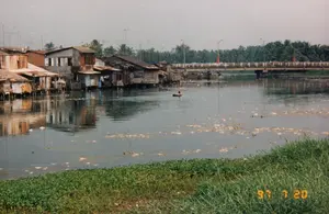 Trên ảnh có ghi ngày tháng '97 7 20'. Bối cảnh là một khu dân cư ven sông hoặc kênh rạch. Dọc theo bờ sông là một dãy nhà sàn, nhà tạm được dựng trên các cọc gỗ ngay trên mặt nước. Các ngôi nhà này được làm từ những vật liệu đơn giản như gỗ, ván và mái tôn. Trên mái nhà có thể thấy các cột ăng-ten truyền hình. Trên mặt sông có rác và lục bình trôi nổi. Một người đang ngồi trong một vật nổi trông giống như chiếc phao (ruột xe) và di chuyển trên sông. Ở phía xa, một cây cầu bê tông bắc qua sông, trên cầu có người đi lại và cắm vài lá cờ đỏ. Dưới gầm cầu có vài chiếc ghe, tàu đang neo đậu. Bờ sông phía xa là những hàng cây dừa và cây cối rậm rạp. Tiền cảnh là bờ cỏ xanh.。（日本語訳）。写真には「'97 7 20」という日付が書かれています。背景は川または運河沿いの住宅地です。川岸に沿って、水面上に直接木の杭で建てられた高床式の家や仮設住宅が並んでいます。これらの家は、木材、板、トタン屋根などの簡素な材料で作られています。屋根の上にはテレビアンテナの柱が見えます。川面にはゴミやホテイアオイが浮かんでいます。一人の人が浮き輪（タイヤチューブ）のような浮遊物に乗って川を移動しています。遠くには、川に架かるコンクリートの橋があり、橋の上には人々が往来し、いくつかの赤い旗が立てられています。橋の下には、数隻の小舟や船が停泊しています。遠くの川岸には、ココナッツの木々や鬱蒼とした木々が並んでいます。前景は緑の草の岸です。