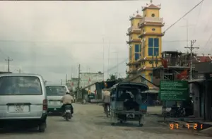 Bức ảnh chụp một con đường đất tại một khu vực có thể là Trà Vinh hoặc Cần Thơ vào ngày 21 tháng 7 năm 1997. Con đường chưa được trải nhựa, và hai bên là những ngôi nhà dân và cửa hàng san sát nhau. Bầu trời u ám.。Nổi bật ở phía bên phải là một công trình kiến trúc lớn màu vàng với hai ngọn tháp cao, có kiểu dáng giống cổng vào của một ngôi chùa Khmer, một nét đặc trưng của vùng Đồng bằng sông Cửu Long. Bên cạnh công trình này là một tòa nhà khác đang trong quá trình xây dựng, để lộ ra phần giàn giáo. Dọc theo các ngôi nhà, nhiều lá cờ đỏ sao vàng của Việt Nam được treo lên, và có rất nhiều cột điện, dây điện cùng ăng-ten TV kiểu "xương cá" chằng chịt, một hình ảnh phổ biến của thời kỳ này.。Trên đường có sự pha trộn giữa các loại phương tiện. Ở phía trước bên trái là một chiếc xe hơi 7 chỗ hiệu Toyota màu bạc. Một người đàn ông đang điều khiển xe máy đi phía trước. Một chiếc xe lam (xe ba gác máy) màu xanh, một phương tiện giao thông công cộng phổ biến thời đó, đang chở nhiều hành khách. Phía sau, một người đội nón lá đang đi xe đạp.。Hoạt động chính là giao thông và sinh hoạt hàng ngày của người dân. Bên lề đường có một tấm biển lớn màu xanh lá cây, chữ trên đó đã mờ nhưng có thể nhận ra đây là biển thông tin dự án xây dựng, với các từ tiếng Anh như "Project", "Employer", "Contractor", "Consultant" và có thể liên quan đến một dự án cấp nước ("Provincial Water Supply Project").。（日本語訳）。1997年7月21日に、おそらくチャーヴィン省かカントー市の地域で撮影された未舗装の道路の写真です。道路はまだ舗装されておらず、両脇には民家や商店が密集しています。空は曇っています。右側で目立っているのは、メコンデルタ地方の特徴であるクメール寺院の門のような様式をした、2つの高い塔を持つ大きな黄色の建造物です。この建造物の隣には、建設中の別の建物があり、足場が見えています。家々に沿って、ベトナムの金星紅旗が多く掲げられており、たくさんの電柱、電線、そしてこの時代によく見られた「魚の骨」型のテレビアンテナが張り巡らされています。道路には様々な種類の乗り物が混在しています。左手前には、銀色のトヨタ製7人乗り自動車があります。一人の男性が前方をバイクで走っています。当時一般的な公共交通機関であった青いラム（三輪自動車）が、多くの乗客を乗せています。その後ろでは、ノンラーをかぶった人が自転車に乗っています。主な活動は、人々の交通と日常生活です。道端には大きな緑色の看板があり、文字は薄れていますが、「Project」、「Employer」、「Contractor」、「Consultant」といった英単語があり、おそらく給水プロジェクト（「Provincial Water Supply Project」）に関連する建設プロジェクトの情報看板であることがわかります。