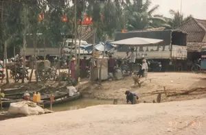 写真は、1997年7月21日、農村地域、おそらくメコンデルタの運河沿いにある船着き場または小さな市場での活気ある生活風景を記録したものである。主な活動は、陸路と水路の間での商品の売買と中継である。多くの人々が、傘やシートで覆われた臨時の屋台の周りに集まっている。運河には数隻の木造の小舟やサンパンがあり、一人の男性がそのうちの一隻に座っている。岸には、おそらく農産物が入っているであろう大きな白い袋がたくさんある。輸送手段には、2台の大きなトラック、多くのバイク（いくつかは大きな竹かごを積んでいる）、1台の自転車、そして1台の手押し車が含まれる。背景には、瓦屋根の家々と、カユプテやヤシの木のような生い茂る木々がある。遠くに赤い看板が一つあるが、文字は読むことができない