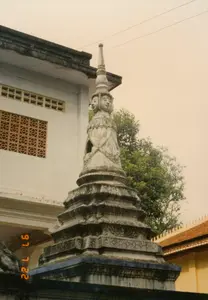 Bức ảnh chụp một ngôi bảo tháp bằng đá trong khuôn viên của một ngôi chùa Khmer. Bảo tháp có kiến trúc nhiều tầng, phần đế được chạm khắc hoa văn tinh xảo đã nhuốm màu thời gian. Phần thân trên của bảo tháp là một bức tượng có bốn khuôn mặt nhìn ra bốn hướng, một đặc điểm của nghệ thuật điêu khắc Khmer. Phía sau bên trái là một tòa nhà bê tông với các ô cửa thông gió bằng gạch hoa. Phía sau bên phải là cây xanh và một phần của một tòa nhà khác có tường vàng và mái ngói kiểu truyền thống. Khung cảnh tĩnh lặng, không có hoạt động của con người. Ở góc dưới bên trái ảnh có dấu ngày tháng màu cam ghi '97 7 22', cho biết ảnh được chụp vào ngày 22 tháng 7 năm 1997.。（日本語訳）。写真は、クメール寺院の境内にある石造りの仏塔を写したものです。仏塔は多層構造で、基壇には精巧な文様が彫刻されており、時の流れを感じさせます。仏塔の上部には、四方を向いた四つの顔を持つ像があり、これはクメール彫刻芸術の特徴です。左後方には、透かしブロックの通気口があるコンクリートの建物があります。右後方には、緑の木々と、黄色い壁と伝統的な瓦屋根を持つ別の建物の一部が見えます。景色は静かで、人の活動は見られません。写真の左下隅には「'97 7 22」と記されたオレンジ色のタイムスタンプがあり、写真が1997年7月22日に撮影されたことを示しています。