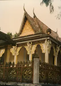 Bức ảnh chụp một công trình kiến trúc tôn giáo, rất có thể là một ngôi chùa Khmer ở Sóc Trăng, thể hiện qua phong cách kiến trúc đặc trưng. Ngôi chùa có mái ngói nhiều tầng, các góc mái cong vút lên tạo thành hình đầu đao, một nét điển hình của chùa Khmer Nam Bộ. Phần trán tường hình tam giác dưới mái được trang trí bằng các hoa văn chạm nổi màu vàng công phu. Hành lang bên ngoài có các cột trụ được trang trí bằng những bức phù điêu tinh xảo, có thể là hình tượng tiên nữ Apsara hoặc chim thần Garuda. Phía trước công trình là một hàng rào được điêu khắc tỉ mỉ với họa tiết trung tâm là hình Bánh xe Pháp luân (Dharmachakra), một biểu tượng quan trọng của Phật giáo. Tổng thể công trình có màu trắng và vàng chủ đạo, đã có dấu hiệu cũ kỹ và rêu phong theo thời gian, phù hợp với bối cảnh năm 1997. Không có hoạt động nào của con người diễn ra và không có văn bản nào có thể đọc được trong ảnh.。（日本語訳）。この写真は宗教建築物を写したもので、その特徴的な建築様式から、ソクチャン省にあるクメール寺院である可能性が非常に高い。寺院は多層の瓦屋根を持ち、屋根の角は鋭く反り上がって「ダウダオ」の形を成しており、これは南部クメール寺院の典型的な特徴である。屋根の下にある三角形の破風は、精巧な金色の浮き彫り模様で装飾されている。外側の回廊には柱があり、天女アプサラまたは神鳥ガルーダと思われる精巧なレリーフで装飾されている。建物の正面には、仏教の重要なシンボルである法輪（ダルマチャクラ）の形を中心的なモチーフとして細かく彫刻された柵がある。建物全体は白と金を基調としており、経年による古びた様子や苔むした痕跡が見られ、1997年という時代背景と一致している。写真には人の活動は見られず、読み取れる文字もない。