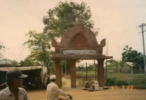 Bức ảnh chụp một con đường đất ở vùng nông thôn, nổi bật là một chiếc cổng lớn mang phong cách kiến trúc đặc trưng của người Khmer. Trên cổng có một tấm biển với dòng chữ Khmer, có thể là tên của một ngôi chùa hoặc một phum sóc (làng của người Khmer). Dòng chữ được xác định là "វត្តមហាសាលវ័ន" (Wat Mahasalavan).。Về hoạt động, một người đàn ông đội mũ lưỡi trai trắng đang điều khiển xe máy trên con đường đất. Gần cổng, hai người phụ nữ đội nón lá đang ngồi nghỉ bên cạnh những chiếc quang gánh và giỏ tre đan. Ở phía trước bên trái, một người đàn ông khác đội mũ lưỡi trai đang quan sát khung cảnh.。Các vật thể trong ảnh bao gồm chiếc cổng kiến trúc Khmer, xe máy, nón lá, mũ lưỡi trai, quang gánh, và các giỏ đựng hàng. Bên trái cổng là một căn nhà hoặc quán nhỏ lợp mái lá/tôn. Phía sau là cây cối, hàng rào, cột điện và những ngôi nhà khác, tạo nên một khung cảnh làng quê điển hình.。（日本語訳）。写真は田舎の未舗装の道を写しており、クメール人の特徴的な建築様式の大きな門が際立っている。門の上にはクメール語の文字が書かれた看板があり、それは寺院の名前か、あるいはプム・ソック（クメール人の村）の名前かもしれない。その文字は「វត្តមហាសាលវ័ន」（ワット・マハサラヴァン）であると特定された。活動については、白い野球帽をかぶった男性が未舗装の道でバイクを運転している。門の近くでは、ノンラーをかぶった二人の女性が、天秤棒と竹で編んだかごのそばで座って休んでいる。左手前では、野球帽をかぶった別の男性が景色を眺めている。写真の中の物体には、クメール建築の門、バイク、ノンラー、野球帽、天秤棒、そして商品を運ぶためのかごが含まれる。門の左側には、葉やトタンで葺かれた家または小さな店がある。背景には木々、柵、電柱、そして他の家々があり、典型的な田舎の風景を作り出している。