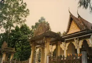 Bức ảnh chụp một ngôi chùa Khmer ở Sóc Trăng, với kiến trúc đặc trưng và tinh xảo. Dấu thời gian kỹ thuật số '97 7 22' được nhìn thấy ở góc dưới bên phải. Ngôi chùa nổi bật với phần mái ngói nhiều tầng, có các đầu đao cong vút, một đặc điểm của kiến trúc chùa tháp Khmer. Các bức tường, đầu hồi và cột trụ được trang trí bằng nhiều hoa văn, phù điêu chạm khắc công phu với tông màu chủ đạo là vàng và trắng. Ở phía trước là một cổng tam quan có mái che, cũng được trang trí cầu kỳ, đồng bộ với kiến trúc tổng thể. Bao quanh khuôn viên là hàng rào được thiết kế với các họa tiết phức tạp. Phía sau và xung quanh chùa là những hàng cây xanh um tùm. Không có hoạt động nào của con người được ghi lại, khung cảnh mang vẻ trang nghiêm và tĩnh lặng.。（日本語訳）。ソクチャン省にあるクメール寺院を撮影した写真で、特徴的で精巧な建築様式が見られます。右下隅に「97 7 22」というデジタルタイムスタンプが見えます。この寺院は、クメールの仏塔建築の特徴である、反り上がった屋根の端を持つ多層の瓦屋根が際立っています。壁、破風、柱は、金と白を基調とした多くの模様や精巧に彫刻されたレリーフで装飾されています。正面には屋根付きの三門があり、これもまた全体の建築様式と調和して凝った装飾が施されています。境内は複雑な模様でデザインされた柵で囲まれています。寺院の後ろと周りには、青々と茂る木々が並んでいます。人の活動は記録されておらず、その光景は荘厳で静寂な雰囲気を醸し出しています。