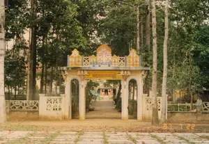 Bức ảnh chụp cổng vào của một ngôi chùa Khmer ở Sóc Trăng, được chụp vào ngày 22 tháng 7 năm 1997. Cổng chùa được xây dựng công phu theo kiến trúc truyền thống của người Khmer, với ba lối đi hình vòm và được sơn màu vàng kem. Phần đỉnh cổng được trang trí lộng lẫy với các họa tiết, phù điêu và các tháp nhỏ nhiều màu sắc. Trên dải băng ngang ở giữa cổng có một dòng chữ Khmer màu vàng trên nền đỏ, nhưng không thể đọc rõ nội dung. Khuôn viên chùa rợp bóng cây cổ thụ. Con đường đất qua cổng dẫn vào sâu bên trong, nơi có thể thấy lờ mờ một công trình khác và một vài người đang đi lại. Sân trước cổng có nhiều lá cây khô rụng trên nền đất và những viên gạch lát đường. Hai bên cổng là hàng rào thấp bằng xi măng có hoa văn trang trí. Khung cảnh mang vẻ yên bình, cổ kính đặc trưng của một cơ sở tôn giáo.。（日本語訳）。ソクチャン省にあるクメール寺院の入口の門を撮影した写真で、1997年7月22日に撮影された。寺院の門はクメール人の伝統建築様式に沿って精巧に建てられており、3つのアーチ型の通路があり、クリームイエローに塗られている。門の頂上部分は、模様、浮き彫り、そして色とりどりの小塔で豪華に装飾されている。門の中央にある横長の帯には、赤地に黄色のクメール文字が一列あるが、内容ははっきりと読み取れない。寺院の境内は古い木々の木陰に覆われている。門の前の庭には、地面にたくさんの枯れ葉が落ちており、敷石もある。門の両側には、装飾的な模様が施されたセメント製の低い塀がある。その光景は、宗教施設特有の平和で古風な趣を醸し出している。