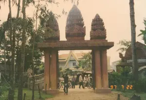 Bức ảnh chụp một cổng tam quan lớn mang đậm kiến trúc Khmer, có thể là lối vào của một ngôi chùa ở Sóc Trăng. Cổng được xây dựng kiên cố với bốn cột trụ lớn, phía trên là ba ngọn tháp được chạm khắc hoa văn công phu. Con đường phía trước cổng là đường đất. Bên trong cổng, có một vài người dân và xe máy. Một người đàn ông mặc áo sẫm màu đang đi bộ ra ngoài. Một nhóm người khác, trong đó có một người đội nón lá, đang đứng gần những chiếc xe máy. Phía sau họ, có thể nhìn thấy một phần của một tòa nhà khác với mái ngói màu vàng và tường xanh, có thể là chánh điện hoặc một công trình khác trong khuôn viên chùa. Cảnh vật xung quanh có nhiều cây xanh, đặc biệt là hàng cây bạch đàn cao ở bên trái. Ở góc dưới bên phải của ảnh có dấu ngày tháng màu vàng ghi "97 7 22". Ở phía xa bên phải, có một cấu trúc cổng khác có chữ viết, nhưng không thể đọc được, dường như là chữ Khmer.。（日本語訳）。写真は、クメール建築様式が色濃く反映された大きな三関門を写しており、おそらくソクチャン省にある寺院の入り口であろう。門は4本の大きな柱で頑丈に建てられており、上部には精巧な文様が彫刻された3つの塔がある。門の前の道は未舗装路である。門の内側には、数人の地元の人々とバイクがいる。暗い色の服を着た一人の男性が、外へ歩いて出ている。別のグループの人々が、その中にはノンラー（菅笠）をかぶった人もおり、バイクの近くに立っている。彼らの後ろには、黄色い瓦屋根と青い壁を持つ別の建物の⼀部が見え、本堂か、あるいは寺院の境内にある別の建造物であろう。周囲の景色には緑の木々が多く、特に左側には背の高いユーカリの木が並んでいる。写真の右下隅には、「97 7 22」と記された黄色い日付の印がある。右側の遠方には、文字が書かれた別の門のような構造物があるが、読むことはできず、クメール文字のようである。