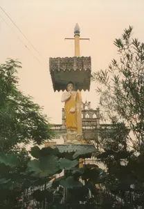 Bức ảnh chụp một bức tượng Phật được đặt ngoài trời, có thể là trong khuôn viên của một ngôi chùa Khmer. Tượng Phật trong tư thế đứng trên một đài sen, khoác áo cà sa màu vàng. Phía trên tượng là một chiếc lọng che được chạm khắc hoa văn tinh xảo. Vươn cao hơn cả là một cây cột, trên đỉnh có trang trí hình đầu tượng nhiều mặt, một đặc điểm thường thấy trong nghệ thuật Khmer. Phía trước tượng là một hồ sen với những chiếc lá lớn màu xanh đậm và vài bông hoa sen màu hồng đang nở. Xung quanh có nhiều cây cối tươi tốt. Phía sau bức tượng là một phần của một công trình kiến trúc lớn, có lẽ là chánh điện hoặc một tòa nhà khác của ngôi chùa, với lan can và các cửa sổ hình vòm. Bầu trời có màu vàng nhạt. Không có chữ viết nào có thể nhận dạng trong ảnh.。（日本語訳）。この写真は屋外に置かれた仏像を写したもので、おそらくクメール寺院の境内にあるものでしょう。仏像は蓮の台座の上に立つ姿で、黄色の袈裟をまとっています。像の上には、精巧な文様が彫刻された天蓋があります。さらにその上には一本の柱が伸びており、その頂上にはクメール美術によく見られる特徴である多面の仏頭像が飾られています。像の前には蓮の池があり、濃い緑色の大きな葉と、咲いているいくつかのピンク色の蓮の花が見えます。周囲には青々とした木々がたくさんあります。像の後ろには、おそらく本堂か寺院の別の建物と思われる大きな建築物の一部があり、手すりやアーチ型の窓が見えます。空は薄い黄色です。写真の中には、識別できる文字はありません。