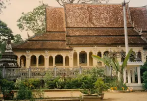 Chữ viết và số: Ở góc dưới bên phải của hình ảnh có một dấu ngày tháng màu vàng cam ghi "97 7 22".。Cảnh quan và hoạt động: Bức ảnh chụp khuôn viên của một ngôi chùa Khmer cổ kính, có khả năng ở Sóc Trăng, dựa trên kiến trúc đặc trưng. Tòa nhà chính, có thể là chánh điện hoặc nhà tăng, được sơn màu vàng nhạt với mái ngói đỏ nhiều tầng đã cũ và lốm đốm. Các chi tiết kiến trúc như hàng cột, cửa vòm, và các họa tiết trang trí trên đầu mái và lan can đều mang đậm phong cách Khmer. Bên trái có một ngôi bảo tháp bằng đá đã phủ rêu phong, cho thấy dấu ấn của thời gian. Phía trước là một khoảng sân đất và một khu vườn nhỏ trồng nhiều loại cây cảnh, bao gồm cả cây chuối quạt. Khung cảnh tĩnh lặng, không có hoạt động nào của con người được ghi lại, tạo cảm giác thanh bình tại một nơi thờ tự.。（日本語訳）。文字と数字：画像の右下隅に、「97 7 22」と記されたオレンジがかった黄色の日付印があります。風景と活動：この写真は、特徴的な建築様式から、おそらくソクチャン省にある古風なクメール寺院の境内を撮影したものです。本堂か僧坊と思われる主要な建物は、薄い黄色に塗られ、古びてまだらになった多層の赤い瓦屋根を持っています。柱の列、アーチ型の扉、屋根の先端や手すりの装飾模様といった建築の細部は、すべてクメール様式を色濃く反映しています。左側には苔むした石造りの仏塔があり、時の経過の跡を示しています。手前には土の庭と、旅人の木を含む多くの種類の観賞植物が植えられた小さな庭園があります。静かな光景で、人間の活動は記録されておらず、礼拝の場所における平和な感覚を生み出しています。