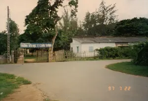 Trên tấm biển hiệu màu xanh phía trên cổng trường có ghi: Dòng trên: PHÒNG GIÁO DỤC HUYỆN CHÂU THÀNH. Dòng dưới: TRƯỜNG TIỂU HỌC T.T CHÂU THÀNH.。Bối cảnh là một con đường nhựa ở vùng nông thôn hoặc thị trấn, dẫn đến cổng của một trường tiểu học. Ngôi trường là một tòa nhà một tầng, lợp mái ngói xi măng, tường sơn trắng, trông khá cũ. Cổng trường được xây bằng hai trụ bê tông với cánh cổng bằng gỗ đã cũ kỹ, một bên dường như đã bị hỏng và được dựng tạm. Xung quanh trường có rất nhiều cây xanh, bao gồm một cây cổ thụ lớn ngay cạnh cổng. Bên trái có một cột điện bê tông. Khung cảnh vắng lặng, không có người hay phương tiện qua lại. Dấu ngày tháng "97 7 22" được in ở góc dưới bên phải của bức ảnh.。（日本語訳）。校門の上にある青い看板には次のように書かれている：上の行：チャウタイン県教育室。下の行：チャウタイン町立小学校。背景は、田舎または町の舗装された道で、小学校の門に通じている。学校の周りにはたくさんの緑の木があり、門のすぐ隣には大きな古木が1本含まれている。左側にはコンクリートの電柱が1本ある。風景は静かで、人や乗り物の往来はない。写真の右下隅に「97 7 22」という日付が印刷されている。