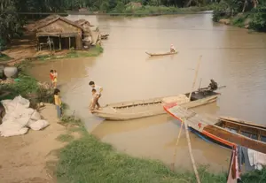 この写真は、農村地域の川沿いの日常の生活風景を記録したもので、ソクチャン省の地域である可能性があります。川の水は、沖積土の濁った茶色をしています。川岸には、葉で葺いた壁の簡素な高床式の家があります。その近くには、水を貯めるためのいくつかの大きな陶器の甕と、土手に積まれた白い麻袋の山があります。水辺の近くで遊んでいる子供たちが何人かいます。一人の女の子が立って見ており、もう一人の子が小さい子を抱いています。川では、一隻のモーター付きボート（エビの尾のようなエンジン付きボート）が岸に着こうとしています。一人の男性が、子供がボートに乗り込むのを手伝っています。ボートの上にも、いくつかの麻袋が積まれています。遠くの方では、一人の人が一人で小さな手漕ぎボートを漕いでいます。右下の角には、洗濯物を干すために使われている別のボートの一部が見えます。主な活動は、ベトナム南西部の水郷地帯に特徴的な、水路による生活と移動です