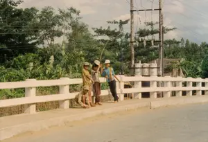 Bức ảnh chụp một nhóm bốn đứa trẻ đang đứng chơi trên một cây cầu bê tông ở một vùng nông thôn. Các em đều đi chân đất và mặc quần áo đơn giản. Ba trong số bốn em đội mũ lưỡi trai. Một em đang ngồi xổm trên lề đường của cây cầu, trong khi ba em còn lại đứng, trong đó có một em đang tựa vào lan can. Một em đang cầm một cành cây dài, có thể là một chiếc cần câu tự chế. Cây cầu có lan can bằng bê tông sơn trắng. Phía sau các em là khung cảnh làng quê với cây cối xanh tươi, rậm rạp, có thể nhận ra các bụi chuối. Phía sau hàng cây có một ngôi nhà mái lá. Hệ thống cột điện và dây điện chằng chịt cũng hiện diện trong khung cảnh.。（日本語訳）。子供たちは皆裸足で、シンプルな服を着ています。4人のうち3人は野球帽をかぶっています。1人は橋の縁にしゃがんでおり、残りの3人は立っていて、そのうちの1人は手すりにもたれかかっています。1人の子供が長い木の枝を持っており、それは手作りの釣り竿かもしれません。橋には白く塗られたコンクリート製の手すりがあります。子供たちの後ろには、青々とした木々が鬱蒼と茂る田舎の風景が広がっており、バナナの茂みが見て取れます。木々の後ろには、茅葺き屋根の家が一軒あります。入り組んだ電柱と電線も風景の中に存在しています。