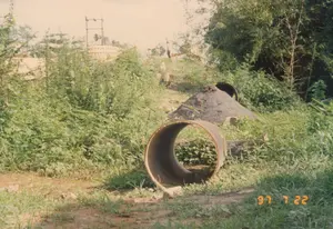 Bức ảnh chụp một khu đất trống, có vẻ như bị bỏ hoang hoặc là một phần của công trường xây dựng ở khu vực nông thôn. Góc dưới bên phải ảnh có dấu ngày tháng màu vàng '97 7 22'. Ở tiền cảnh, một đoạn ống cống lớn bằng bê tông nằm trên mặt đất đầy cỏ dại. Phía sau ống cống là một đống vật liệu tối màu, có thể là cát hoặc sỏi, với một mảnh lốp xe cũ đặt trên đỉnh. Cảnh vật xung quanh um tùm cây cỏ và bụi rậm. Ở phía sau, trên một con dốc, có thể thấy lờ mờ bóng dáng của một vài người, một cột điện, và một công trình kiến trúc có mái vòm thấp ẩn hiện sau lùm cây.。（日本語訳）。写真は空き地を写しており、放棄されているか、あるいは田舎の建設現場の一部のようです。写真の右下隅には、「'97 7 22」という黄色い日付スタンプがあります。前景には、大きなコンクリート製の土管が雑草の生い茂る地面に置かれています。土管の後ろには、暗い色の資材の山があり、おそらく砂か砂利で、その上には古いタイヤの破片が置かれています。周囲の景色は草木や茂みで生い茂っています。後方、坂の上には、数人の人影、電柱、そして低いドーム型の屋根を持つ建造物が木々の後ろに見え隠れしているのがぼんやりと見えます。