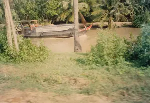 Bối cảnh là một con kênh ở vùng nông thôn, có thể là ở khu vực Đồng bằng sông Cửu Long. Một chiếc ghe lớn, có vẻ là ghe chở hàng, đang di chuyển trên dòng nước màu nâu phù sa. Chiếc ghe này có phần mái che đơn sơ bằng gỗ và khoang hàng được phủ bạt. Bên hông ghe có treo những chiếc lốp xe cũ để chống va đập. Một người đang ở trong khoang lái. Đi bên cạnh là một chiếc xuồng nhỏ hơn, với phần mũi được trang trí màu đỏ và trắng, trên xuồng cũng có một người ngồi. Hoạt động chính là di chuyển và vận chuyển bằng đường thủy. Khung cảnh hai bên bờ kênh rậm rạp cây cối, nổi bật là những hàng dừa và các loại cây nhiệt đới khác. Không có văn bản hay chữ viết nào có thể nhận dạng được trong ảnh.。（日本語訳）。背景は田舎の運河で、おそらくメコンデルタ地域でしょう。大きな船、見たところ貨物船のようですが、それが沖積土で茶色くなった水の上を移動しています。この船には木製の簡素な屋根があり、貨物室は防水シートで覆われています。船の側面には、衝撃を防ぐために古いタイヤが吊るされています。一人の人が操縦室にいます。その隣にはより小さなボートが並走しており、船首は赤と白で装飾され、そのボートにも一人の人が乗っています。主な活動は水路による移動と輸送です。運河の両岸の景色は木々が鬱蒼としており、特にヤシの木やその他の熱帯植物の列が目立ちます。写真の中には、認識できるテキストや文字はありません。