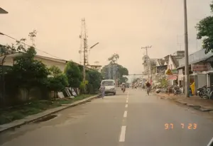 Bản ghi chữ viết: Có một số biển hiệu có thể đọc được. Một biển hiệu lớn màu đỏ và trắng ghi "LE MINH TAM". Một biển hiệu khác ghi "NHÀ THUỐC TÂY". Ngoài ra còn có một biển báo giao thông hình tam giác màu vàng, viền đỏ, cảnh báo về giao lộ phía trước.。Mô tả cảnh quan và hoạt động: Bức ảnh chụp một con đường nhựa ở một thị trấn hoặc khu vực ngoại ô tại Việt Nam, vào ngày 23 tháng 7 năm 1997. Khung cảnh cho thấy hoạt động giao thông thường ngày với nhiều người đi xe đạp và xe máy. Một chiếc xe tải nhỏ (van) màu trắng đang ở bên trái đường, gần đó có một người phụ nữ mặc đồ bộ đang đi bộ. Bên phải đường là một dãy nhà phố liền kề, được sử dụng làm cửa hàng, có nhiều xe đạp và xe máy đậu trên vỉa hè. Bầu trời có nhiều mây. Cơ sở hạ tầng nổi bật với rất nhiều cột điện, dây điện và dây cáp chằng chịt trên không, cùng với một tháp viễn thông lớn ở phía xa bên trái. Lề đường bên trái có cỏ mọc và một bức tường.。（日本語訳）。書かれている文字：読み取れる看板がいくつかある。赤と白の大きな看板には「LE MINH TAM」と書かれている。別の看板には「NHÀ THUỐC TÂY」と書かれている。その他に、前方の交差点を警告する、赤枠の黄色い三角形の交通標識がある。風景と活動の描写：写真は1997年7月23日、ベトナムの町または郊外地域にある舗装道路を写したものである。光景は、多くの人々が自転車やバイクで行き交う日常の交通の様子を示している。道路の左側には白い小型トラック（バン）がおり、その近くをセットアップの服を着た女性が歩いている。道路の右側には、店舗として利用されている長屋風の建物が並んでおり、歩道には多くの自転車やバイクが停められている。空には雲が多い。道路の左側の路肩には草が生え、壁がある。