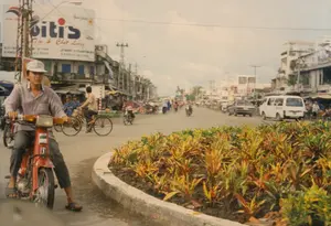 Bức ảnh chụp một con đường phố đông đúc ở Việt Nam vào khoảng năm 1997. Ở tiền cảnh, một người đàn ông đang ngồi trên chiếc xe máy Honda Cub màu đỏ, nhìn về phía máy ảnh. Ông mặc áo sơ mi dài tay, quần tối màu, đi dép và đội mũ lưỡi trai. Con đường có nhiều người và phương tiện qua lại, bao gồm xe máy, xe đạp, một chiếc xe van màu trắng và một chiếc ô tô con, phản ánh cảnh sinh hoạt giao thông thường ngày. Hai bên đường là những dãy nhà phố cao hai đến ba tầng. Hệ thống dây điện và cột điện chằng chịt phía trên. Một tấm biển quảng cáo lớn của thương hiệu Biti's rất nổi bật, trên đó có các dòng chữ: 'Biti's', 'Uy Tín & Chất Lượng', 'Sản phẩm', 'HƯƠNG TƯƠI', và các thông tin liên hệ như 'KC CTY 129 [...] ĐT 8754070', 'CỬA HÀNG TPHCM 88-B 8752443', 'KC CHI NHÁNH [...] ĐT 87521729 FAX 71[...]'. Ở giữa đường là một bồn hoa được xây viền bê tông, trồng các loại cây cảnh có lá nhiều màu sắc.。（日本語訳）。この写真は1997年頃のベトナムの混雑した街路を撮影したものです。前景には、赤いホンダ・カブのバイクに座り、カメラの方を見ている男性がいます。彼は長袖のシャツと暗い色のズボンを着用し、サンダルを履いて野球帽をかぶっています。道路にはバイク、自転車、白いバン、乗用車など、多くの人や乗り物が行き交い、日常の交通風景を反映しています。道路の両側には、2階から3階建ての店舗兼住宅が並んでいます。頭上には電線と電柱が複雑に張り巡らされています。Biti'sブランドの大きな広告看板が非常に目立っており、そこには「Biti's」、「Uy Tín & Chất Lượng（信頼と品質）」、「Sản phẩm（製品）」、「HƯƠNG TƯƠI」といった文言や、「KC CTY 129 [...] ĐT 8754070」、「CỬA HÀNG TPHCM 88-B 8752443」、「KC CHI NHÁNH [...] ĐT 87521729 FAX 71[...]」などの連絡先情報が記載されています。道路の中央にはコンクリートで縁取られた花壇があり、色とりどりの葉を持つ観葉植物が植えられています。
