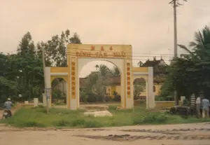 ベトナムのある村の亭の三門を撮影した写真。門には赤いクオック・グーで「ĐÌNH TÂN NGÃI」と書かれ、その上には漢字で「亭新義」（Đình Tân Nghĩa）とある。門は黄色に塗られ、3つのアーチ型の入口を持つ建築様式である。背景は農村地域または郊外である。前景の道は土の道で、まだ舗装されていない。右側には、電線が複雑に絡み合った電柱が一本立っている。人の動きについては、左側で一人の人が道をバイクで走っている。右側の遠くには、数人のグループが立っている。この光景は、1997年頃のベトナムのある地方における、静かな日常生活の一コマを表している