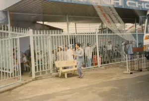 Bối cảnh là lối vào của một cơ sở, có thể là một bến xe khách hoặc một nhà máy, được rào lại bằng một hàng rào và cổng sắt lớn màu trắng. Phía trên cổng có một tấm biển hiệu lớn màu xanh với chữ trắng, nhưng chỉ đọc được một phần '...H CH...'. Một biểu ngữ màu trắng chữ đỏ bị treo ngược, có thể đọc mờ các chữ như 'CHẤT LƯỢNG' và 'T-XN'. Nhiều người, bao gồm cả một người đội nón lá, đang đứng chờ phía sau cổng sắt. Ở phía trước, một người đàn ông mặc bộ đồng phục công nhân màu xám đang đi bộ. Gần cổng có một chiếc ghế đá màu vàng có khắc chữ nhưng không rõ nội dung. Phía bên phải của bức ảnh là một phần đầu của một chiếc xe buýt, và có các rào chắn di động được đặt gần đó. Khung cảnh cho thấy hoạt động chờ đợi để di chuyển hoặc ra vào một khu vực có kiểm soát.。（日本語訳）。背景は、ある施設の入り口で、バスターミナルか工場の可能性があり、大きな白い鉄の柵と門で囲われています。門の上には、白文字の大きな青い看板がありますが、「...H CH...」という部分しか読み取れません。赤い文字の白い横断幕が逆さまに吊るされており、「CHẤT LƯỢNG」や「T-XN」といった文字がぼんやりと読み取れます。ノンラーをかぶった一人を含む多くの人々が、鉄の門の後ろで待っています。手前では、灰色の作業服を着た男性が歩いています。門の近くには、文字が刻まれた黄色い石のベンチがありますが、内容は不明です。写真の右側にはバスの先端部分があり、その近くには移動式のバリケードが設置されています。この光景は、移動するため、あるいは管理された区域に出入りするために待っている様子を示しています。