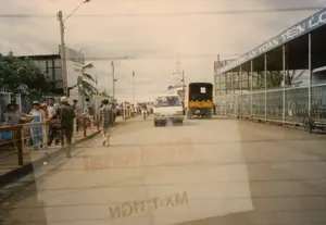 Bản ghi văn bản:。- Trên mái che của tòa nhà bên phải có một biển hiệu lớn màu xanh với dòng chữ trắng: "...NHANH CHÓNG AN TOÀN TIỆN LỢI".。- Trên bức tường trắng của một tòa nhà ở bên trái có chữ màu đỏ nhưng bị mờ và khó đọc.。Mô tả cảnh quan và hoạt động:。- Địa điểm: Cảnh quay trên một con đường nhựa ở Việt Nam, có thể là khu vực gần Sài Gòn hoặc Củ Chi. Con đường có vẻ đông đúc. Bên trái là lối đi dành cho người đi bộ có lan can. Bên phải là một công trình lớn có hàng rào sắt kéo và mái che, trông giống như một bến xe, nhà ga hoặc lối vào một nhà xưởng.。- Hoạt động: Nhiều người dân đang đi bộ trên lối đi bộ. Một người phụ nữ đội nón lá, có thể đang mang hàng hóa. Một người đàn ông mặc áo sơ mi kẻ sọc đang đi bộ dưới lòng đường. Các phương tiện như xe tải nhỏ, xe tải lớn và xe máy đang lưu thông trên đường. Đây là một cảnh sinh hoạt và giao thông thường ngày vào năm 1997.。- Vật thể: Có một chiếc xe tải nhỏ màu trắng và một chiếc xe tải lớn màu vàng đang di chuyển trên đường. Nhiều người đi bộ, trong đó có một phụ nữ đội nón lá. Có các cột điện, dây điện và đèn đường. Một chiếc xe đạp được dựng gần hàng rào sắt bên phải. Bức ảnh được chụp từ bên trong một phương tiện, có hình ảnh phản chiếu của chữ viết trên cửa kính.。（日本語訳）。テキストの記録：。- 右側の建物の庇の上には、「...迅速 安全 便利」と書かれた大きな青い看板がある。- 左側の建物の白い壁には赤い文字があるが、ぼやけていて読みにくい。風景と活動の描写：。- 場所：ベトナムのアスファルト道路での光景で、サイゴンまたはクチ近郊の地域と思われる。左側は手すりのある歩行者用の通路である。- 活動：多くの人々が歩道を歩いている。ノンラーをかぶった女性がおり、商品を運んでいる可能性がある。縞模様のシャツを着た男性が車道を歩いている。小型トラック、大型トラック、バイクなどの車両が道路を通行している。これは1997年における日常生活と交通の光景である。- 物体：白い小型トラック1台と黄色い大型トラック1台が道路を走行している。多くの歩行者がおり、その中にはノンラーをかぶった女性もいる。電柱、電線、街灯がある。右側の鉄柵の近くに自転車が1台立てかけられている。写真は車両の中から撮影されたもので、窓ガラスに文字の反射が写っている。