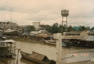 Trên hình ảnh có dấu ngày tháng màu cam: 97 7 23.。Đây là khung cảnh một khu dân cư đông đúc ven một con sông hoặc kênh rạch có màu nước đục ngầu, đặc trưng của vùng sông nước. Bên bờ sông là một khu chợ tạm, nơi nhiều người đang tụ tập mua bán dưới những mái che bằng bạt và mái lá. Hoạt động mua bán diễn ra tấp nập.。Trên sông có nhiều loại thuyền ghe. Ở phía trước là một chiếc ghe nhỏ bằng gỗ có mái che bằng lá. Cập sát bờ chợ là những chiếc tàu lớn hơn, có động cơ, dường như được dùng để vận chuyển hàng hóa hoặc làm nơi buôn bán nổi. Ở góc dưới bên trái có một cấu trúc giống như nhà sàn hoặc một cái cầu tàu bằng gỗ.。Ở hậu cảnh là nhiều ngôi nhà san sát nhau, từ những ngôi nhà lợp tôn, mái lá đơn sơ đến những tòa nhà bê tông kiên cố. Trên nóc các tòa nhà có rất nhiều ăng-ten TV, một hình ảnh phổ biến vào thời điểm đó. Một tháp nước cao bằng bê tông nổi bật trên nền trời. Có thể nhìn thấy một lá cờ đỏ sao vàng của Việt Nam trên một trong những ngôi nhà.。（日本語訳）。画像にはオレンジ色の日付スタンプ「97 7 23」があります。これは、水郷地帯に特徴的な濁った色の川または運河のほとりにある、密集した住宅地の光景です。川岸には仮設の市場があり、多くの人々が防水シートや茅葺きの屋根の下に集まって売買をしています。売買活動は活発に行われています。川には多くの種類の船があります。手前には、茅葺きの屋根が付いた小さな木製の小舟があります。市場の岸に接岸しているのは、より大きなエンジン付きの船で、商品を輸送したり、水上での商売の場として使われたりしているようです。左下隅には、高床式の家または木製の桟橋のような構造物があります。背景には、トタン屋根や茅葺きの簡素な家から頑丈なコンクリートの建物まで、多くの家が密集しています。建物の屋上には多くのテレビアンテナがあり、それは当時の一般的な光景です。コンクリート製の高い給水塔が、空を背景に際立っています。