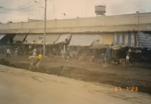 Bức ảnh chụp một khu chợ ven đường vào ngày 23 tháng 7 năm 1997. Ở phía trước, một công nhân đội mũ bảo hộ màu vàng đang dùng xẻng làm việc trên một con đường đất lầy lội, cho thấy có thể đang có công trình xây dựng. Phía sau là một dãy các sạp hàng, quầy hàng được che bằng những tấm bạt tạm, một số có sọc xanh trắng. Nhiều người dân, trong đó có người đội nón lá, đang đi lại và hoạt động mua bán trong khu chợ. Có thể thấy nhiều xe đạp dựng gần các gian hàng. Phía trên và sau khu chợ là một kết cấu bê tông lớn, có thể là một cây cầu vượt hoặc tường của một tòa nhà lớn. Một tháp nước cũng hiện ra ở phía xa trên nóc nhà. Bầu trời có nhiều dây điện và cột điện chằng chịt. Không có văn bản nào có thể đọc được trên các biển hiệu. Dấu ngày tháng "97 7 23" được ghi ở góc dưới bên phải.。（日本語訳）。1997年7月23日に撮影された、ある道端の市場の写真。前景には、黄色いヘルメットをかぶった作業員がぬかるんだ未舗装の道路でシャベルを使って作業しており、建設工事が行われている可能性を示している。後ろには、間に合わせのシートで覆われた露店や屋台が並んでおり、そのうちのいくつかは青と白の縞模様である。ノンラーをかぶった人を含む多くの人々が、市場を行き来し、売買活動をしている。市場の上方と後方には、高架橋か大きな建物の壁と思われる大きなコンクリートの構造物がある。遠くの屋根の上には給水塔も現れている。空には多くの電線と電柱が張り巡らされている。看板には読み取れる文字はない。右下の角に「97 7 23」という日付が記されている。