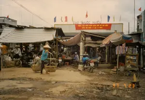 Bối cảnh là một khu chợ trời ở Việt Nam vào ngày 23 tháng 7 năm 1997, có tên là "Chợ Thuộc Nhiêu". Phía trên có một bảng hiệu lớn ghi "CHỢ THUỘC NHIÊU" và một băng rôn màu đỏ với dòng chữ vàng: "NHIỆT LIỆT CHÀO MỪNG NGÀY BẦU CỬ ĐẠI BIỂU QUỐC HỘI KHÓA X (20-7-1997)". Nền chợ là đất, lầy lội và không bằng phẳng. Một người phụ nữ mặc áo xanh, đội nón lá, đang dùng đòn gánh gánh hai chiếc thúng tre lớn. Xung quanh là các gian hàng tạm bợ được che bằng mái bạt sọc và mái tôn. Nhiều người dân đang hoạt động trong chợ, một số ngồi tại các bàn ghế gỗ đơn sơ, có thể là ở một quán ăn nhỏ. Có một vài chiếc xe máy, phương tiện phổ biến thời bấy giờ, đang đậu gần các gian hàng. Một quầy hàng nhỏ bên phải, dưới một chiếc ô lớn, bày bán các mặt hàng như nước đóng chai và thuốc lá trên kệ gỗ. Nhiều cờ phướn đủ màu sắc và cờ đỏ được treo phía trên, tạo không khí cho sự kiện bầu cử.。（日本語訳）。背景は1997年7月23日のベトナムにある「トゥックニエウ市場」という名の屋外市場です。上方には「CHỢ THUỘC NHIÊU」と書かれた大きな看板と、「第10期国会議員選挙の日（1997年7月20日）を熱烈に歓迎します」という黄色い文字が書かれた赤い横断幕があります。市場の地面は土で、ぬかるんでおり、平らではありません。青い服を着てノンラーをかぶった一人の女性が、天秤棒で二つの大きな竹かごを担いでいます。周りには、縞模様の防水シートやトタンの屋根で覆われた仮設の屋台があります。多くの人々が市場で活動しており、何人かは簡素な木製のテーブルと椅子に座っています。そこは小さな食堂かもしれません。当時の一般的な交通手段であった数台のバイクが、屋台の近くに停まっています。右側には大きな傘の下に小さな売店があり、木製の棚にペットボトル飲料やタバコなどの商品を並べて販売しています。上方には色とりどりの多くの幟旗や赤い旗が掲げられており、選挙イベントの雰囲気を醸し出しています。