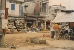 Bối cảnh là một khu chợ ven đường hoặc một con phố ở Việt Nam vào khoảng năm 1997. Con đường bằng đất, chưa được trải nhựa. Dọc hai bên đường là những ngôi nhà tầng kiểu cũ, tầng trệt được sử dụng làm cửa hàng tạp hóa với mái hiên bằng bạt sọc hoặc tôn. Hoạt động chính là buôn bán và sinh hoạt đời thường. Một số người bán hàng, trong đó có những phụ nữ đội nón lá, đang ngồi trên vỉa hè để bán các loại hàng hóa như trái cây được bày trên mâm. Một nhóm đàn ông đang đứng nói chuyện gần những chiếc xe máy đang đậu. Các vật thể đáng chú ý bao gồm những chiếc xe máy kiểu cũ, giỏ tre, và trên một cây cột điện có dán một tờ giấy quảng cáo nhưng chữ viết đã mờ và không thể đọc rõ.。（日本語訳）。背景は1997年頃のベトナムの道端の市場またはある通りです。道は土で、まだ舗装されていません。道の両脇には古い様式の階建ての家が並び、1階は縞模様のシートやトタンの軒先を持つ雑貨店として使われています。主な活動は商売と日常生活です。ノンラーをかぶった女性を含む何人かの売り子が、歩道に座って盆に盛られた果物などの商品を売っています。男性の一団が、停まっているバイクの近くで立ち話をしています。注目すべき物には、古い様式のバイク、竹かご、そして電柱に貼られた広告紙がありますが、文字はかすれていてはっきりと読むことはできません。