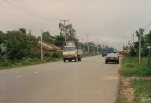 Cảnh quan một con đường quốc lộ ở vùng nông thôn hoặc ngoại ô Việt Nam vào năm 1997. Con đường được trải nhựa với vạch kẻ đường phân làn. Hoạt động chính là giao thông đường bộ với nhiều loại xe cộ. Nổi bật ở phía trước là một chiếc xe tải màu trắng, thùng xe phủ bạt xanh, đang di chuyển. Bên lề phải có một chiếc ô tô con màu đen. Ở phía xa hơn, có thể nhận ra một chiếc xe buýt màu xanh lam và một vài chiếc xe tải khác. Hai bên đường là cây cối um tùm, bao gồm cả cây chuối. Phía sau hàng cây bên trái là một vài ngôi nhà cấp bốn lợp ngói. Hệ thống cột điện và dây điện chằng chịt chạy dọc theo con đường. Bầu trời u ám, nhiều mây. Không có văn bản hoặc chữ viết nào có thể đọc được trên các biển báo hay phương tiện.。（日本語訳）。1997年、ベトナムの田舎または郊外における国道の景観。道路は舗装されており、車線を分ける白線が引かれている。主な活動は道路交通であり、多くの種類の車両が見られる。前方で目立つのは、荷台が青いシートで覆われた一台の白いトラックで、移動中である。右側の路肩には、一台の黒い乗用車がある。さらに遠くには、一台の青いバスと他の数台のトラックが見て取れる。道路の両側には木々が鬱蒼と茂っており、バナナの木も含まれる。左側の並木の後ろには、数軒の瓦葺きの平屋の家がある。電柱と複雑に絡み合った電線が、道路に沿って続いている。空はどんよりとしており、雲が多い。標識や車両には、読み取れる文字や文章はない。