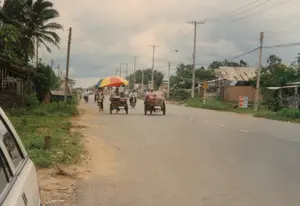 Bức ảnh ghi lại cảnh sinh hoạt trên một con đường ở vùng ngoại ô Việt Nam vào năm 1997, có thể là khu vực Củ Chi hoặc Sài Gòn. Con đường được trải nhựa, hai bên là lề đất và cỏ mọc. Dọc đường là những hàng cột điện với hệ thống dây điện chằng chịt, và những ngôi nhà dân có kiến trúc đơn giản, một số lợp mái tôn.。Hoạt động chính trên đường là giao thông và vận chuyển hàng hóa. Nhiều người đang di chuyển bằng xe đạp. Nổi bật là hai chiếc xe lôi: một chiếc là xe bán hàng rong được đẩy bằng tay, có gắn một chiếc ô lớn sặc sỡ (vàng, đỏ, xanh) để che nắng; chiếc còn lại do xe đạp kéo, đang chở những món hàng cồng kềnh được bọc lại. Ngoài ra còn có một vài người đi xe máy. Ở phía trước bên trái là một phần của một chiếc ô tô.。Cây cối, bao gồm cả cây dừa, mọc xanh tốt hai bên đường. Có một số biển hiệu: một biển báo giao thông hình tam giác màu vàng cảnh báo có người đi bộ hoặc trẻ em; một tấm biển quảng cáo trên tường gạch có chữ 'BÁN TẬN GỐC' nhưng phần còn lại bị mờ; và một tấm biển khác quảng cáo 'DẠY LÁI XE' cùng với các dịch vụ ăn uống như 'CƠM', 'BIA', 'NƯỚC NGỌT'.。（日本語訳）。この写真は1997年、ベトナムの郊外のある道路での生活風景を記録したもので、クチまたはサイゴン地区の可能性があります。道路はアスファルトで舗装されており、両側は土の路肩で草が生えています。道沿いには、電線が複雑に絡み合った電柱が並び、簡素な建築様式の民家が建ち並び、中にはトタン屋根の家もあります。道路上の主な活動は、交通と商品の輸送です。多くの人が自転車で移動しています。目立っているのは2台の荷車です。1台は手押しの屋台で、日よけのために大きなカラフルな（黄色、赤、青）傘が取り付けられています。もう1台は自転車で引かれており、梱包されたかさばる荷物を運んでいます。その他にも、バイクに乗っている人が数人います。左手前には、自動車の一部が見えます。ココナッツの木を含む木々が、道の両脇に青々と茂っています。いくつかの看板があります。歩行者や子供に注意を促す黄色の三角形の交通標識が1つ。レンガの壁には「BÁN TẬN GỐC」と書かれた広告看板がありますが、残りの部分はぼやけています。そしてもう1つの看板は、「DẠY LÁI XE」を宣伝しており、「CƠM」、「BIA」、「NƯỚC NGỌT」といった飲食サービスも一緒に書かれています。